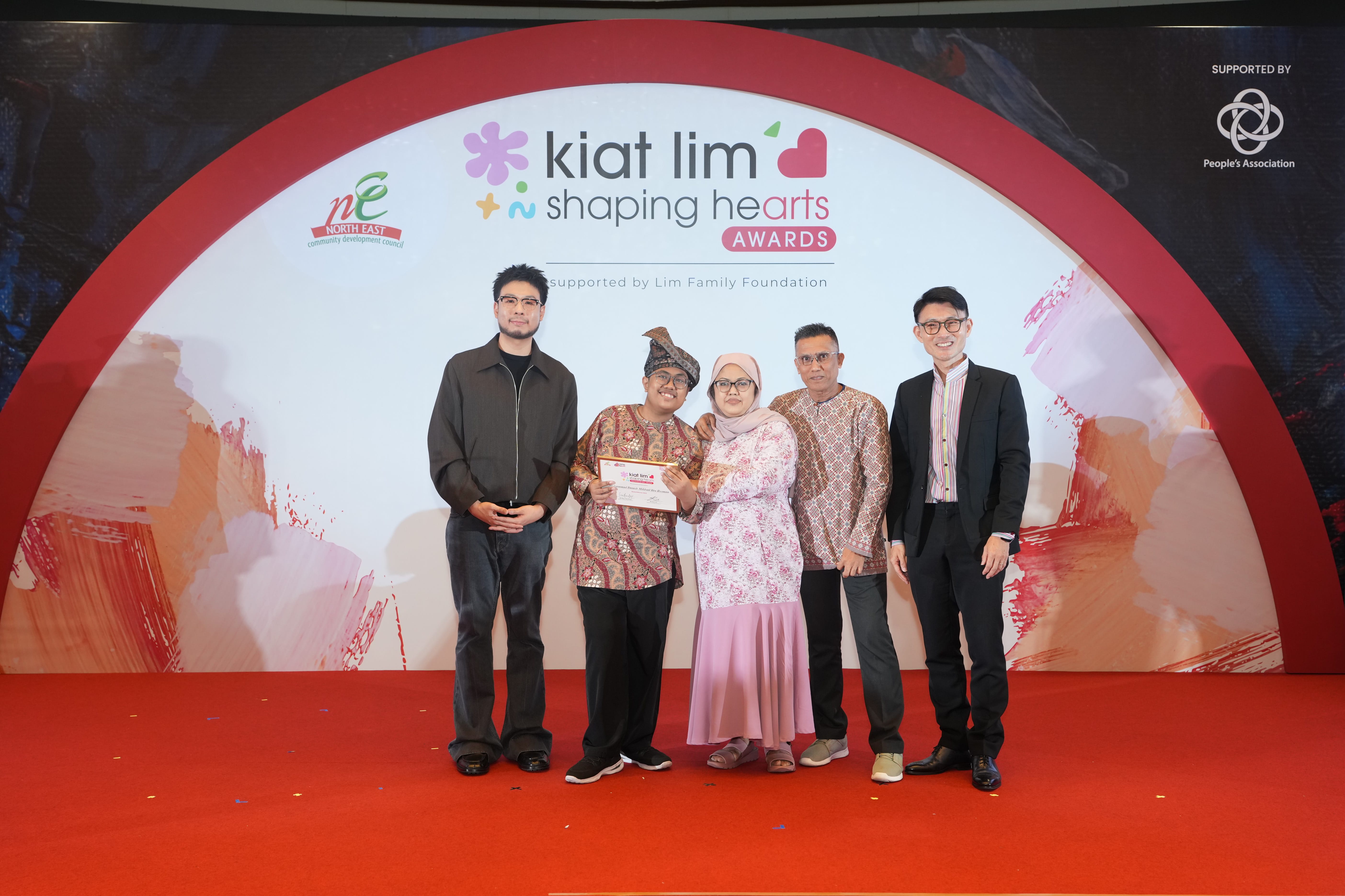 Award recipient standing on stage with the presenter, holding their certificate and posing for a group photo during the award ceremony, with a backdrop reading ‘Kiat Lim-Shaping Hearts Awards'.